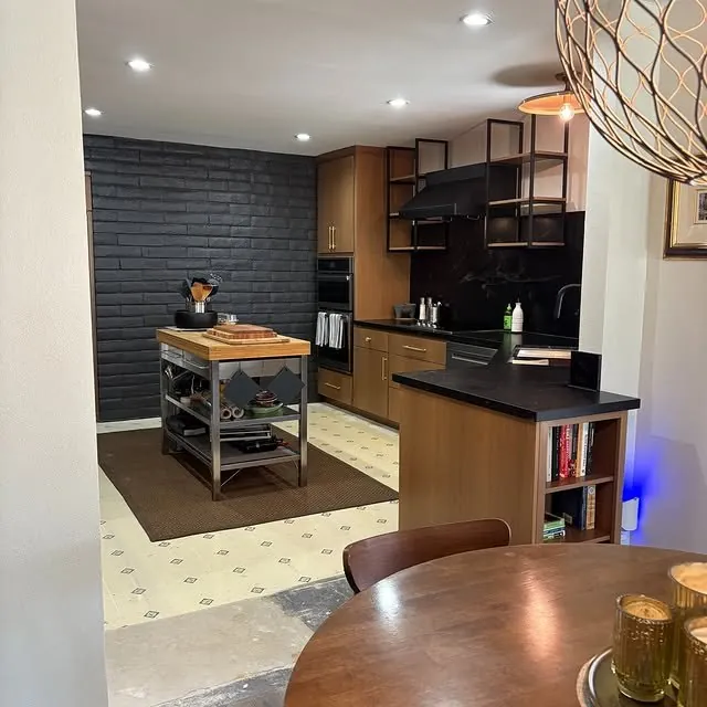 Open kitchen seen from the dining table, with a dark brick wall and a butcher-block cart